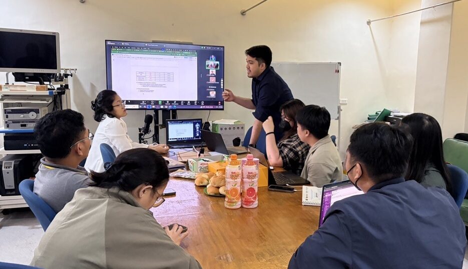 Troyss Pilapil presenting to the V Luna General Hospital team during a meeting, gesturing toward a screen displaying a spreadsheet, while colleagues seated around the table with laptops look on