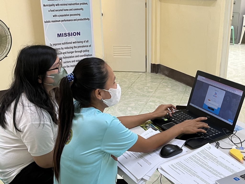 Myrrhkryss presenting a system on a laptop to a barangay health worker (BHW), with the two reviewing the login screen together at a desk covered in paperwork