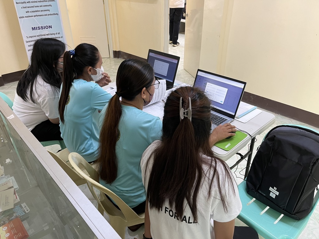 Luisa and Myrrhkryss guiding barangay health workers (BHWs) through the CareGo system on laptops, with the group gathered around a table during a training session.