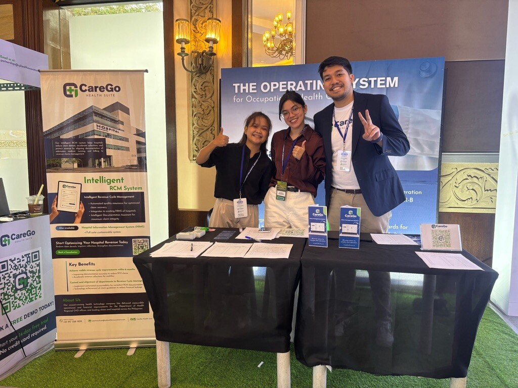 CareGo Health Suite team posing with peace signs and thumbs up at their event booth, with banners promoting their Intelligent RCM System and 'The Operating System for Occupational Health' displayed behind them